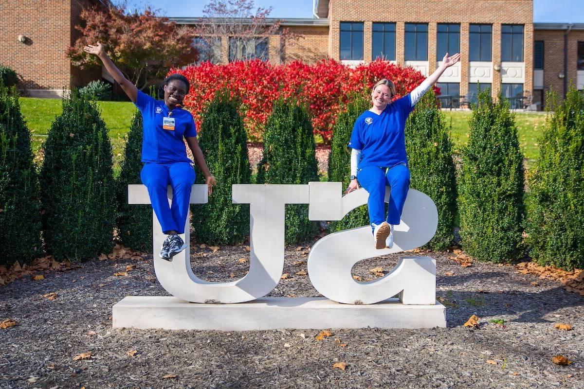 Two Shenandoah nursing students, in blue scrubs, sit atop the SU statue in the Shenandoah University Quad in the autumn.